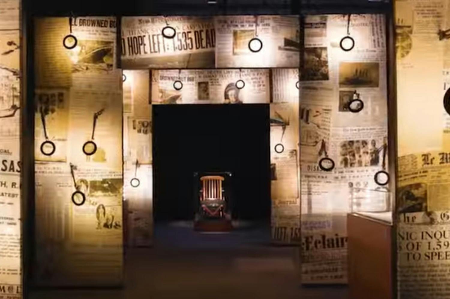 A museum exhibit featuring an electric chair in a darkened space, surrounded by illuminated panels with old newspaper headlines.