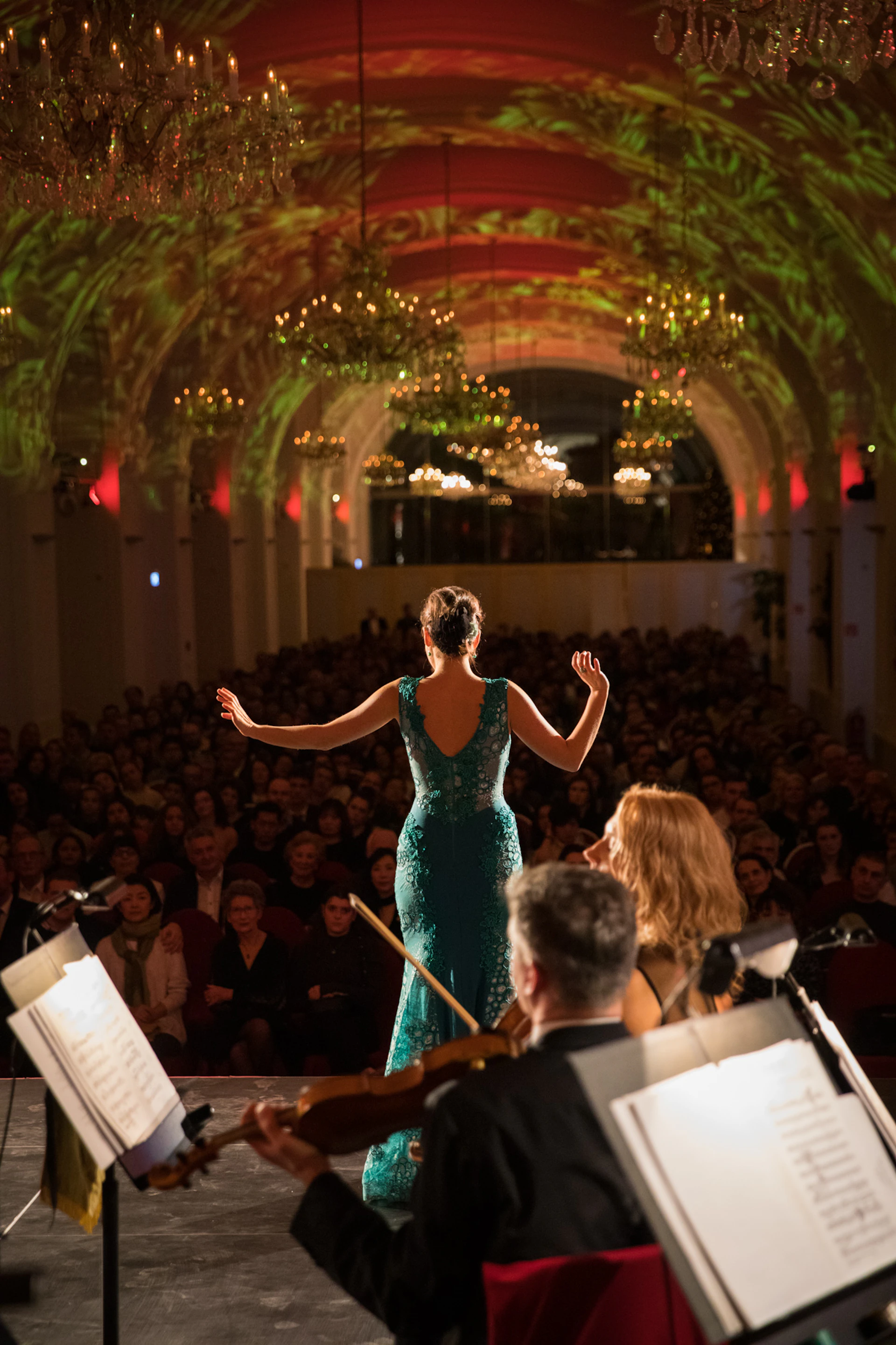 A woman in a green dress performs on stage in a grand hall with chandeliers, facing a large audience.