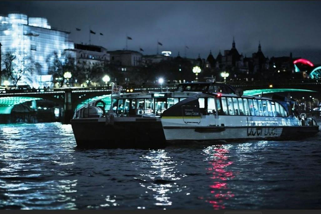 A boat on a river at night with illuminated city buildings and a bridge in the background.