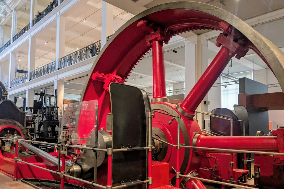 Large red industrial machinery with gears and pipes, displayed in a spacious museum setting.