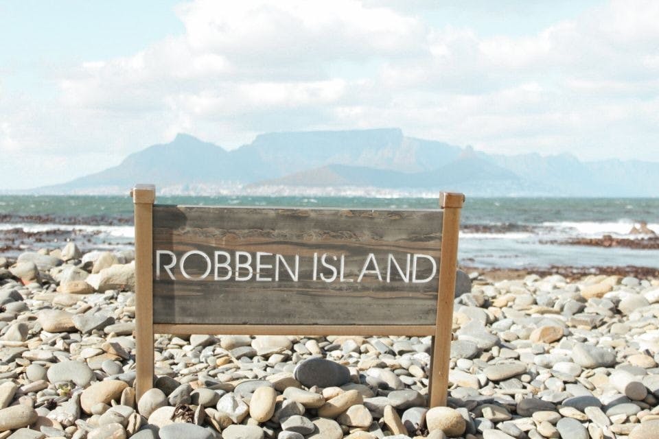Mezi kameny a oblázky stojí dřevěná cedule s nápisem „Robben Island“, v pozadí je vidět oceán a hory.