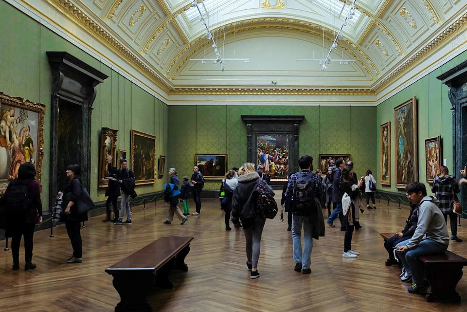 People walking and viewing paintings in a spacious, ornate gallery with green walls and a wooden floor.