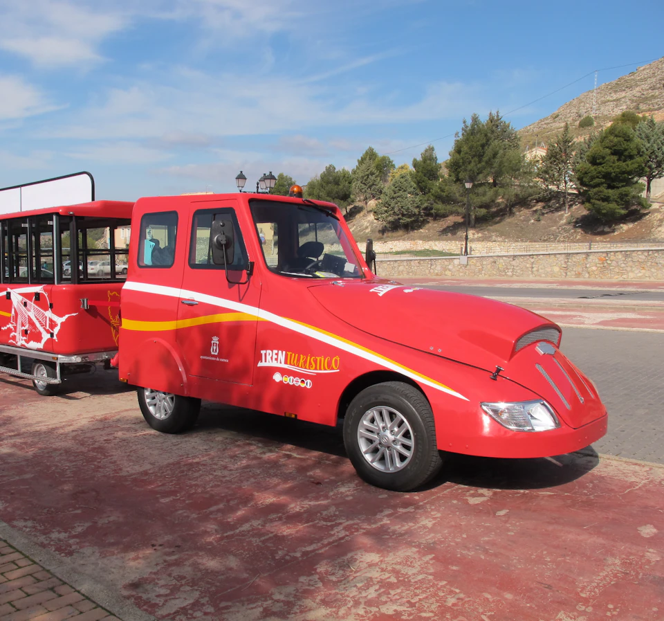A red touristic train with two carriages parked on a paved area, surrounded by trees and hills under a blue sky.