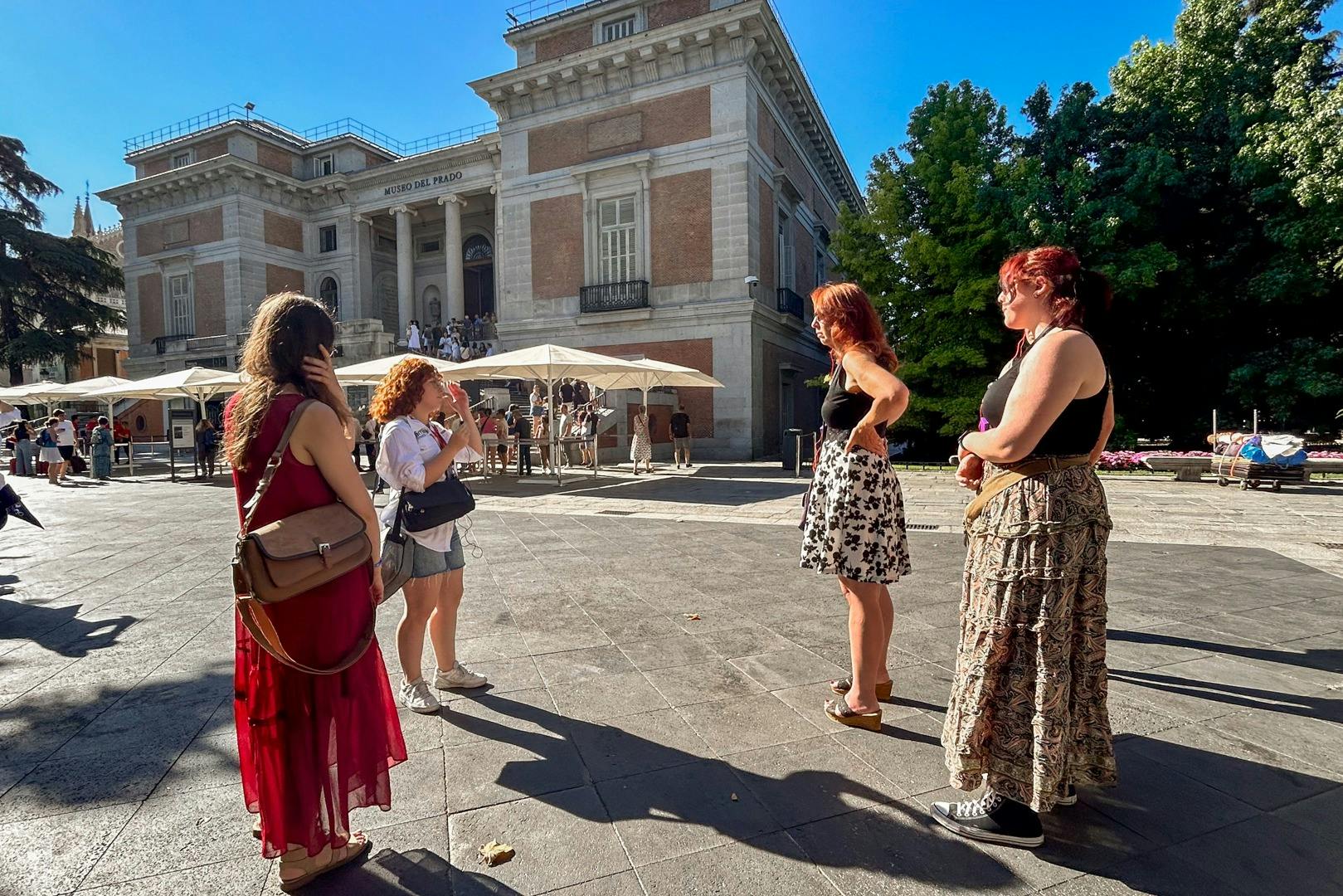벽돌 건물과 나무 근처의 햇살이 내리쬐는 광장에 서 있는 두 여성과 다른 사람들, 우산 아래 앉은 사람들.