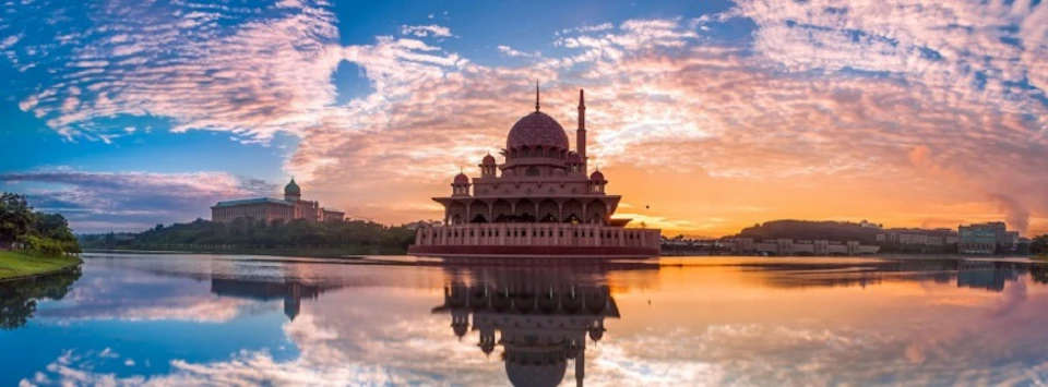 A mosque with a large dome and minaret is reflected in calm water at sunset, with a vibrant sky of orange and blue hues.