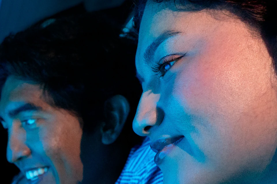 Two people in a submarine look out round windows at underwater scenes, one holding a phone and smiling. Blue lighting fills the space.