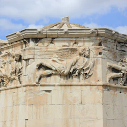 Närbild av en gammal stenstruktur med detaljerade reliefsniderier av bevingade figurer och draperade plagg under en klar himmel.