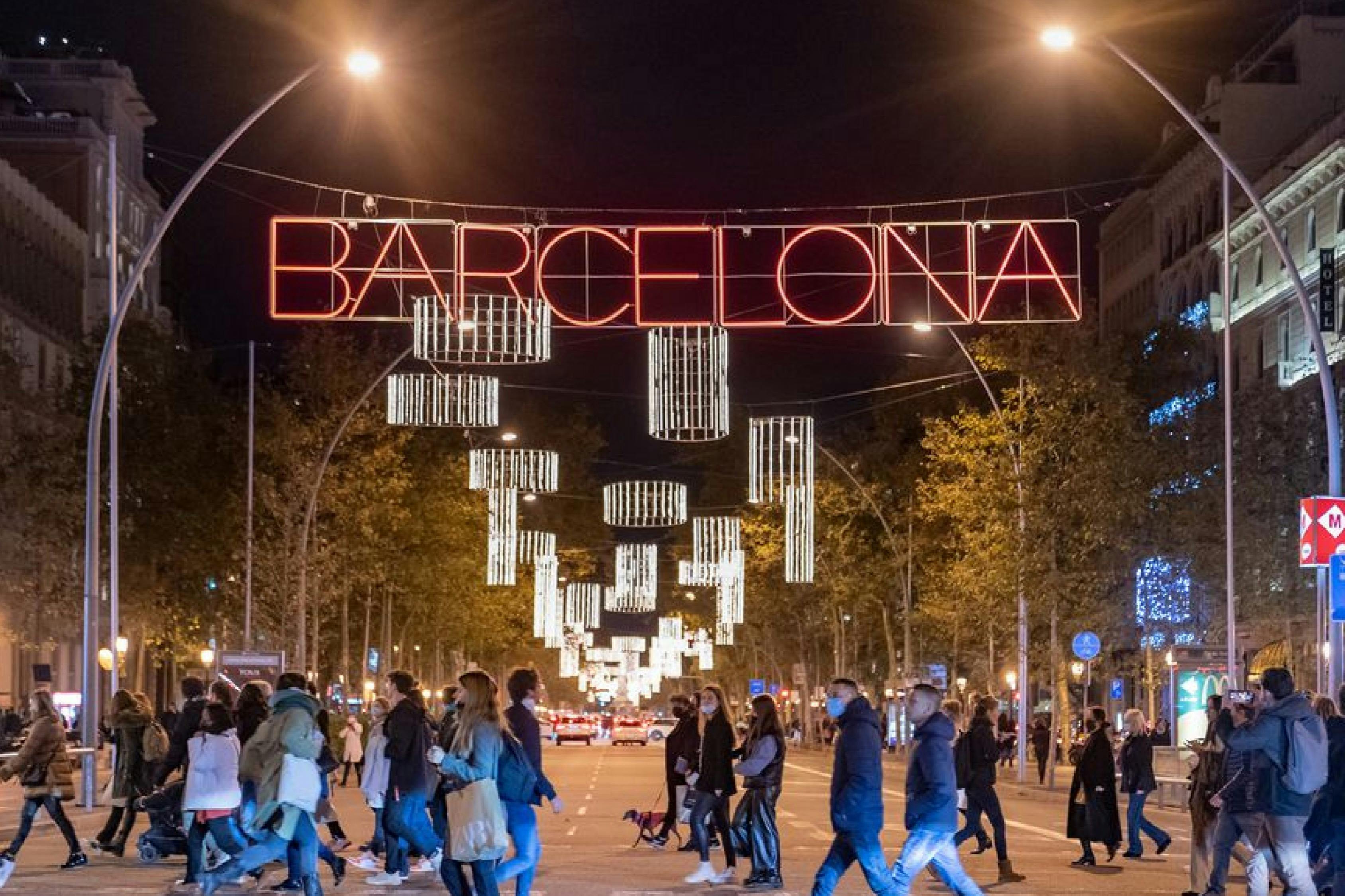 People walking on a street at night with "Barcelona" in red neon lights hanging above and illuminated decorations in the background.