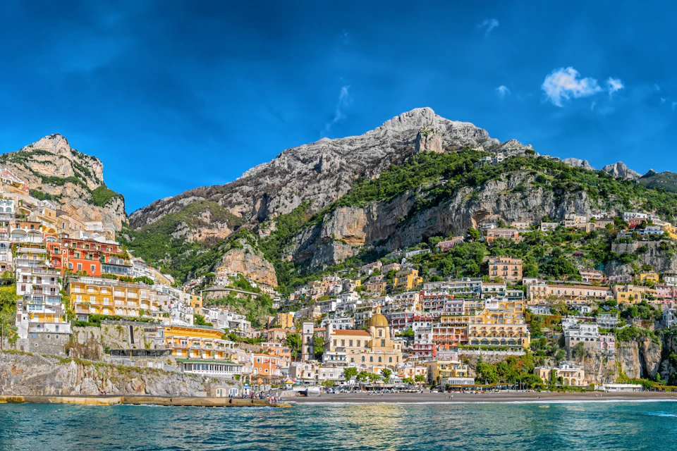 A coastal town with colorful buildings on a steep hillside under a blue sky with a mountainous backdrop.