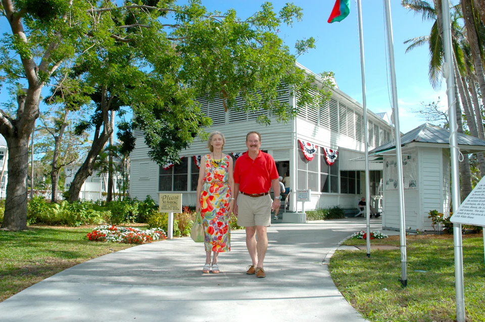 An elderly couple walks on a path outside a white building decorated with patriotic bunting and surrounded by trees and flowers.