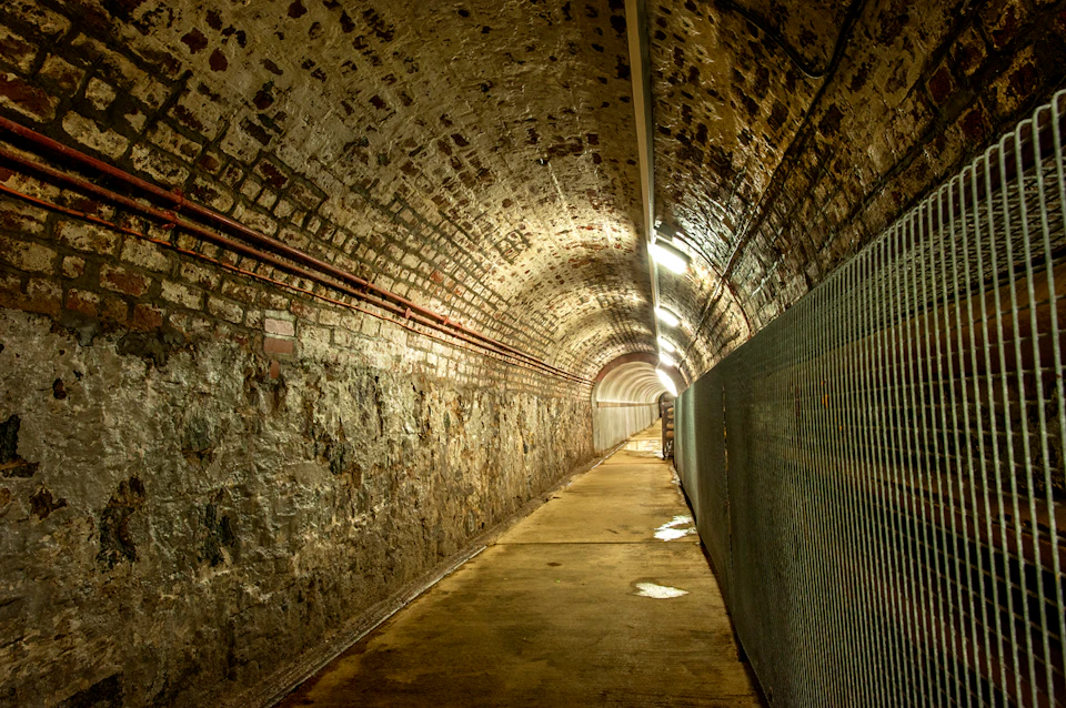 A dimly lit, narrow underground tunnel with brick walls, a curved ceiling, and metal grating on one side.