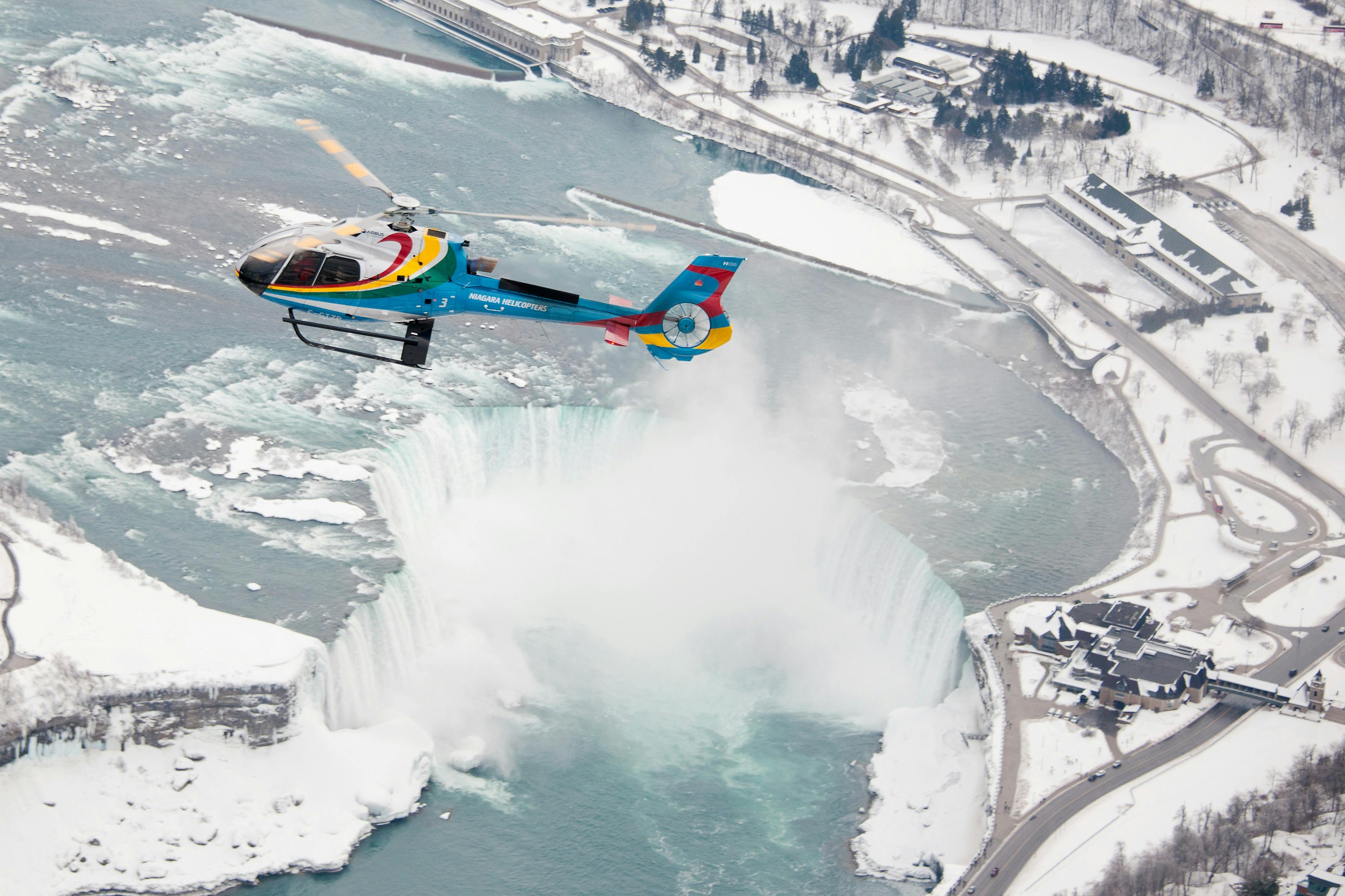 A colorful helicopter flies above Niagara Falls, with snow-covered ground and surrounding buildings visible.