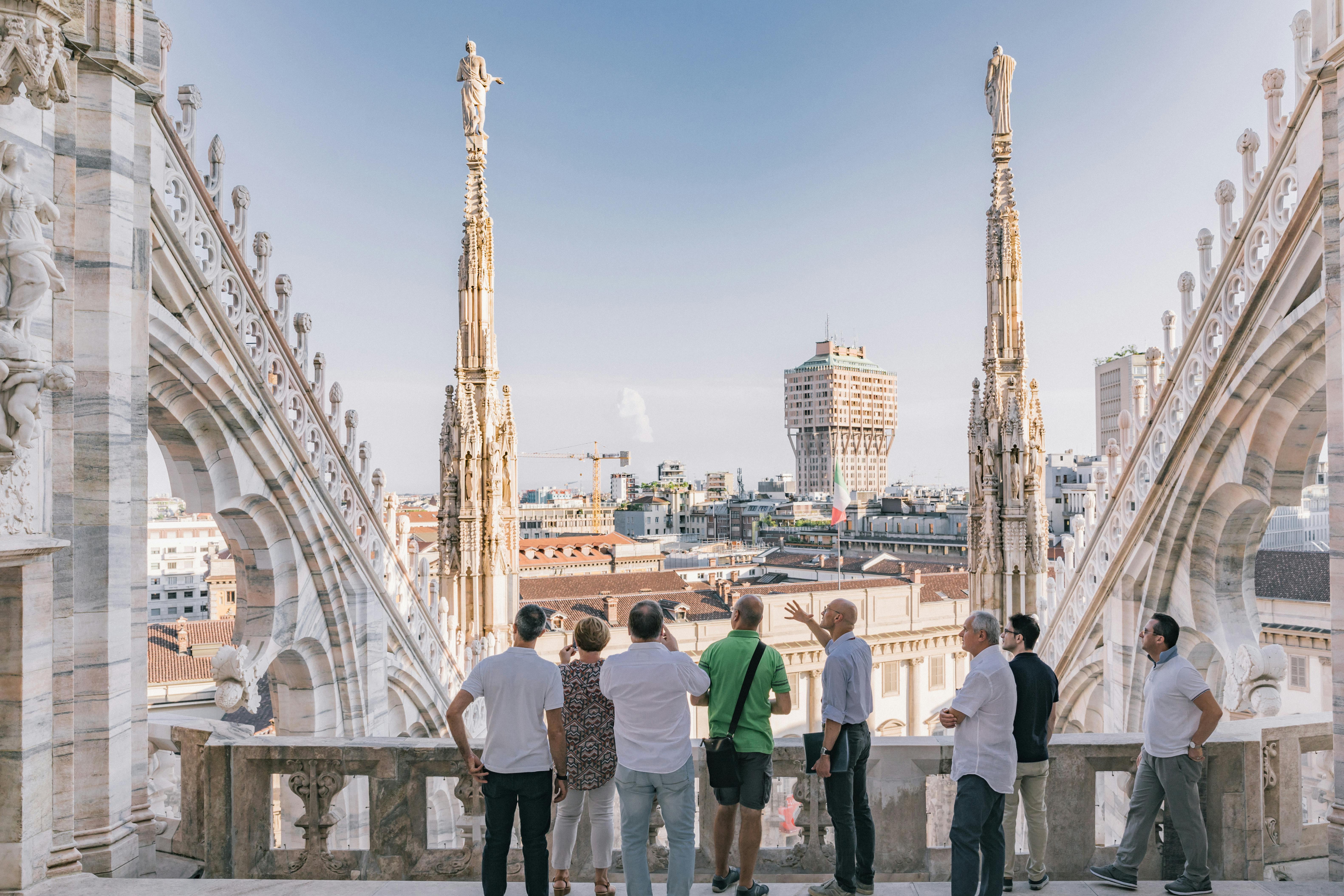 Six people stand on a terrace with ornate pillars, overlooking a cityscape with a distinctive tower in the background under a clear sky.