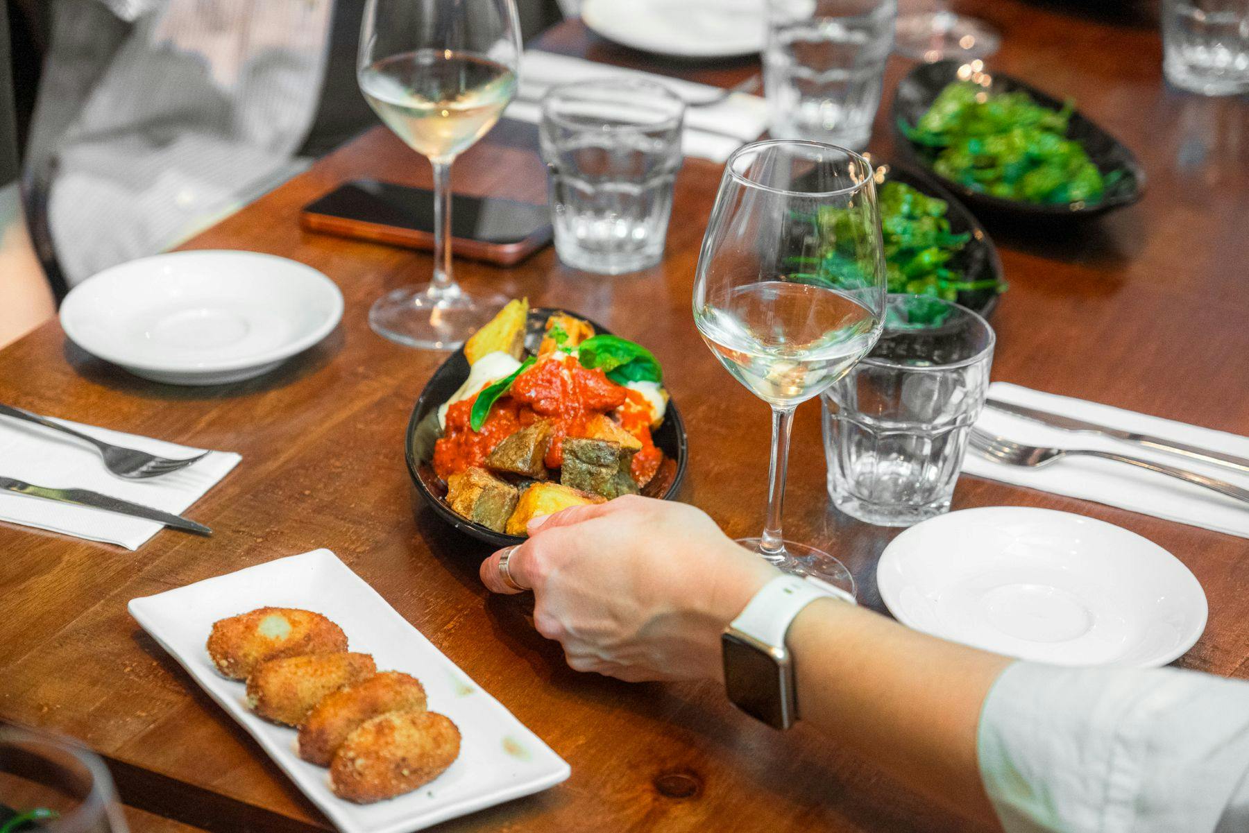 Uma mesa com pratos de comida, copos de vinho e uma mão a pegar num prato, num ambiente de refeição.