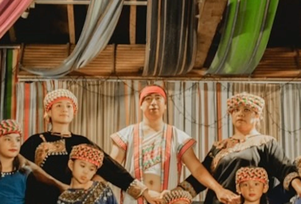 A group of people in traditional attire stand on a stage decorated with colorful fabric, holding hands in a semi-circle.