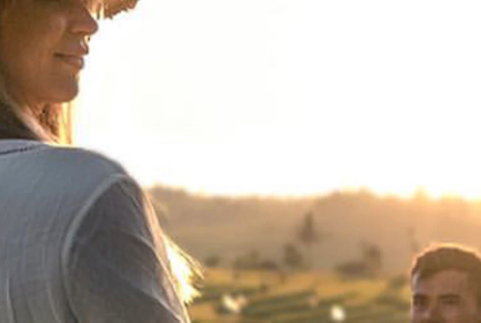 A woman in a white dress and hat stands in a field at sunset, with a man in the background smiling at her.