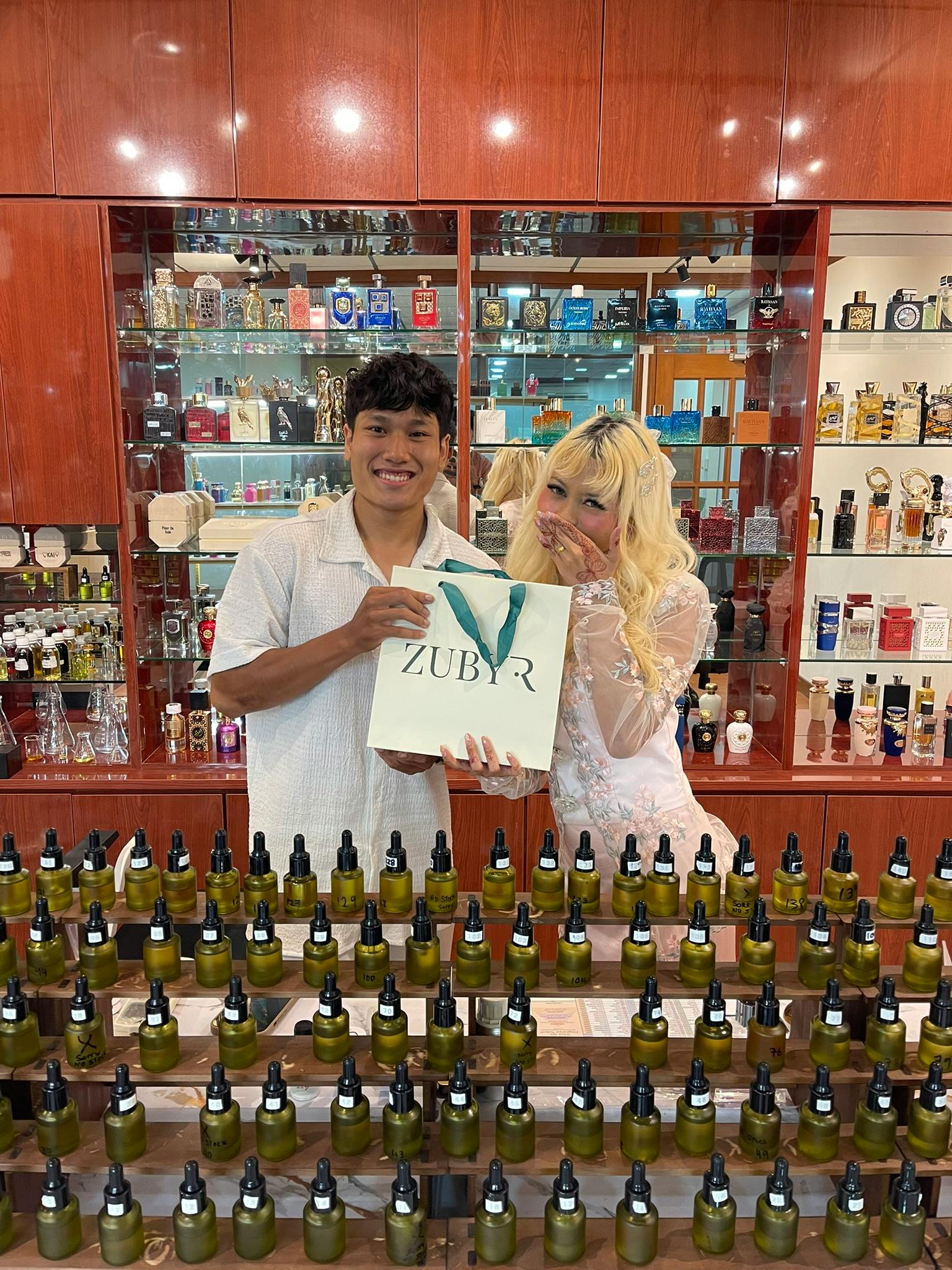 Two people standing in a shop with shelves of perfume bottles, one holding a bag labeled 'ZUBYR,' smiling.