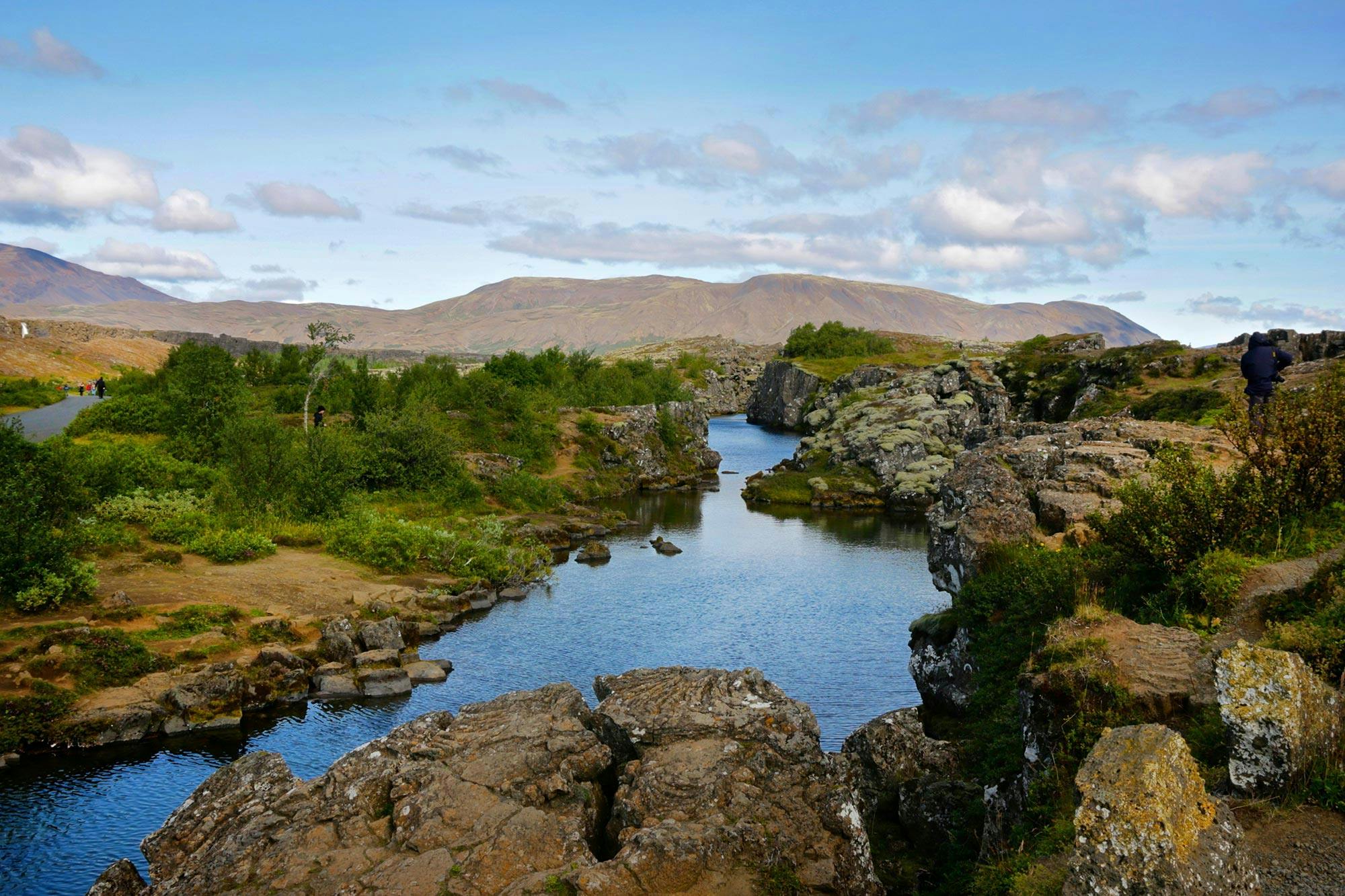Clear river flanked by rocky and grassy terrain, with trees and distant mountains under a blue sky with scattered clouds.