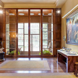 Ett rymligt rum med en stor glasdörr, trägolv, ett konsolbord med dekorativa skålar, en stor inramad målning och en skulptur.