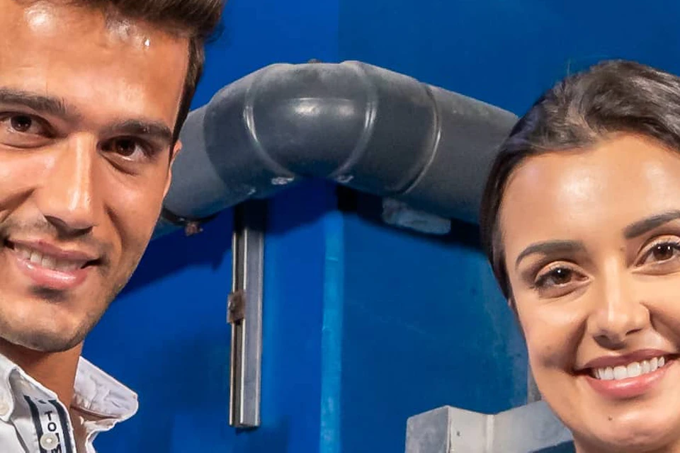 Two people in waders stand in a large tank with several small sharks, blue walls and piping in the background.