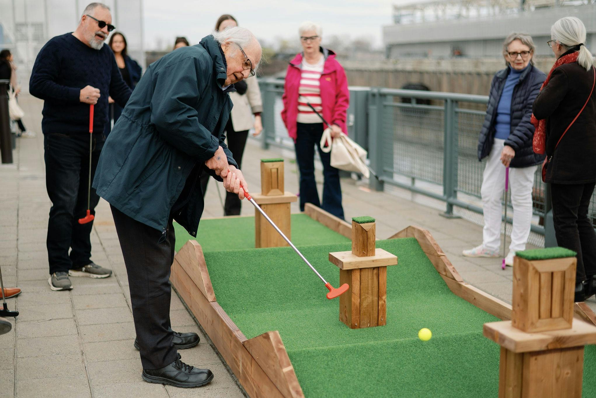 Um homem mais velho a jogar mini-golfe com espectadores que se encontram nas proximidades, numa área pavimentada junto a um gradeamento.