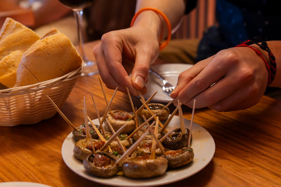 A person places toothpicks in stuffed mushrooms on a plate, with bread and a glass of wine in the background.