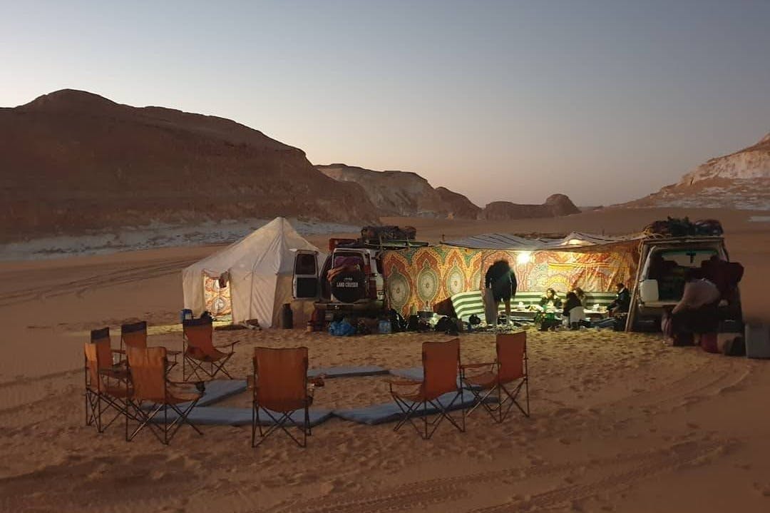 A desert campsite with a white tent, colorful canopy, camping chairs, and a vehicle at sunset. People are seen standing and sitting.