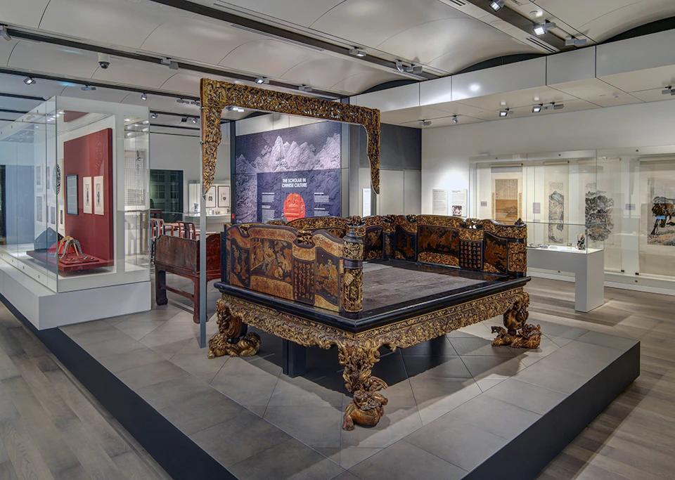 Ornate, antique bed exhibit in a museum with detailed carvings and golden accents, displayed under bright lighting.