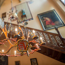 En utsmyckad ljuskrona med ett krön hänger nära en trätrappa dekorerad med invecklade sniderier och inramad konst.