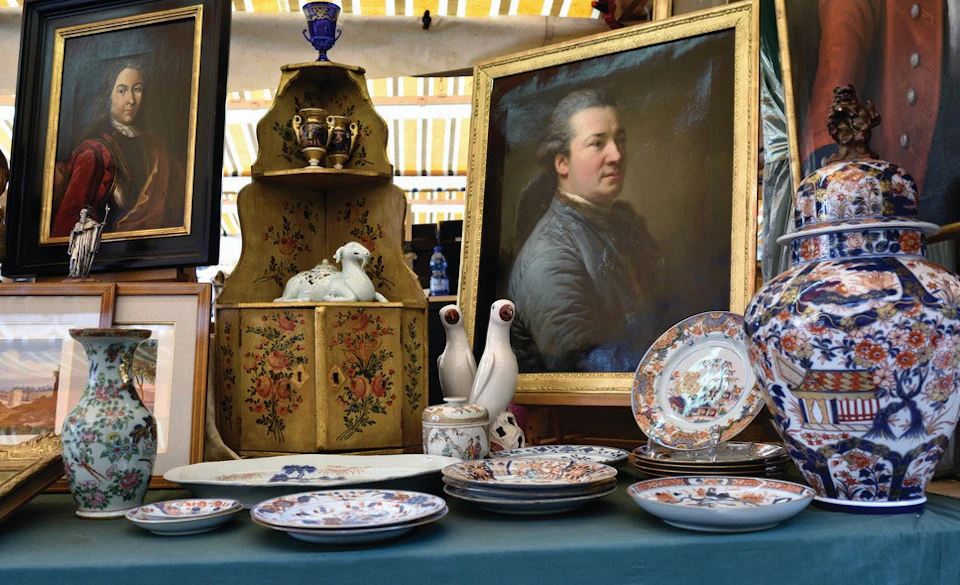 A table displays porcelain plates, vases, and figurines, with a framed portrait of a man in the background.