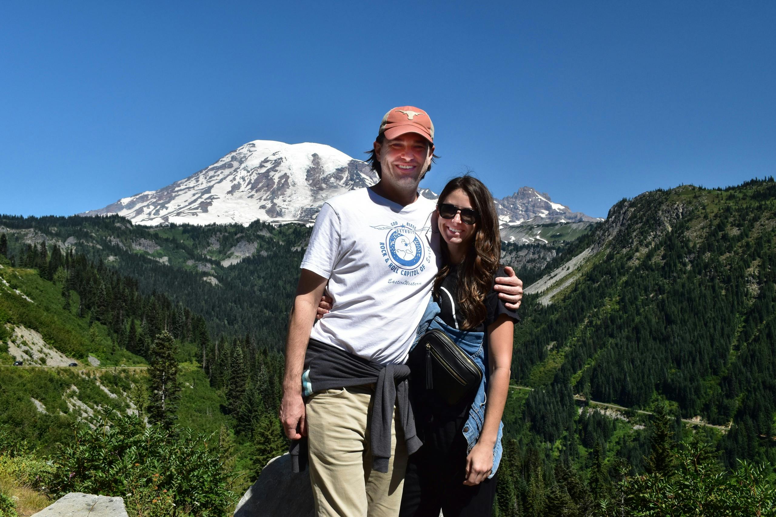 Una coppia in posa all'aperto con una montagna e degli alberi sullo sfondo, sotto un cielo azzurro e limpido.