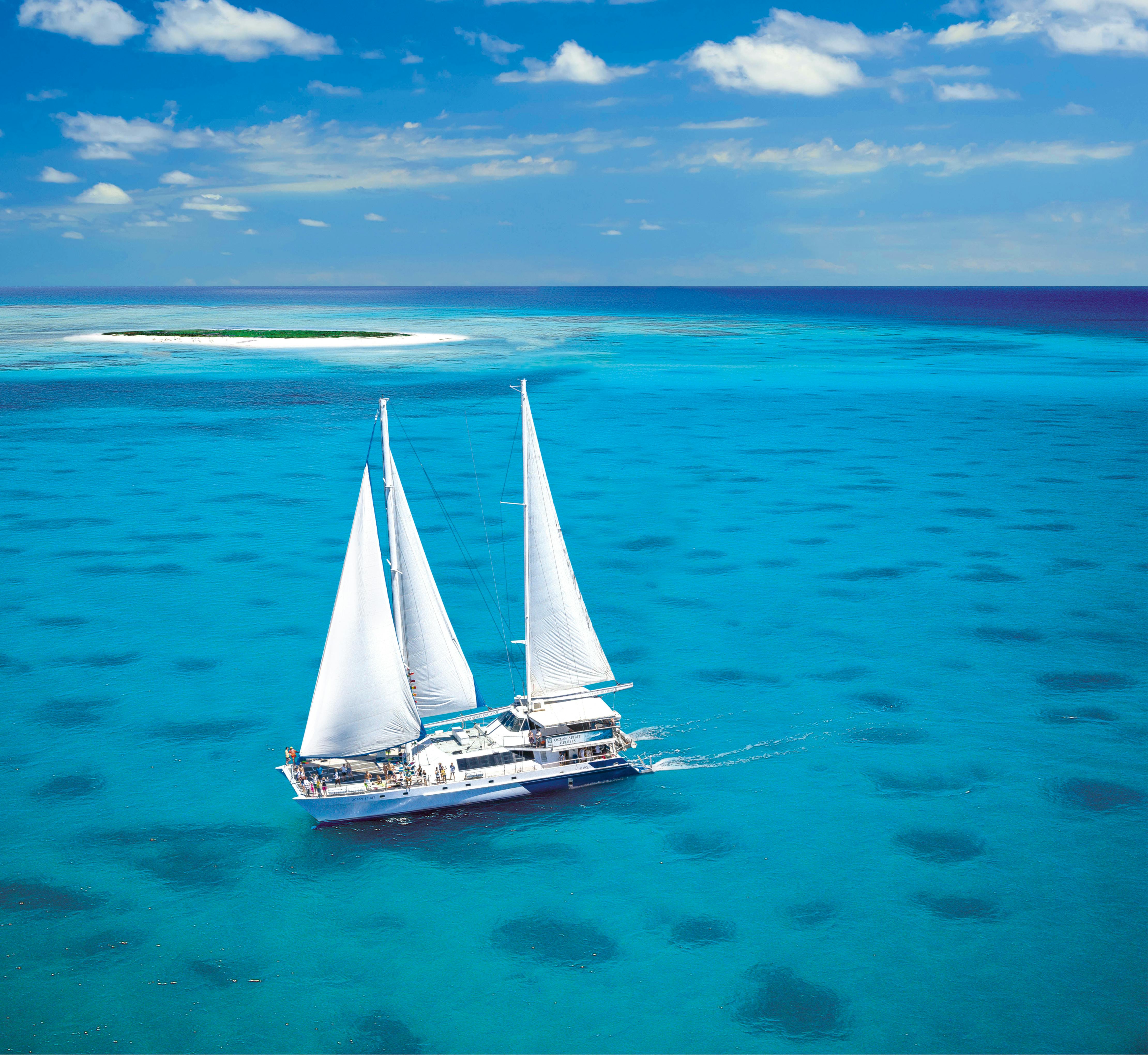 A white sailboat with tall sails glides on clear turquoise water, near a small sandy island under a blue sky with scattered clouds.