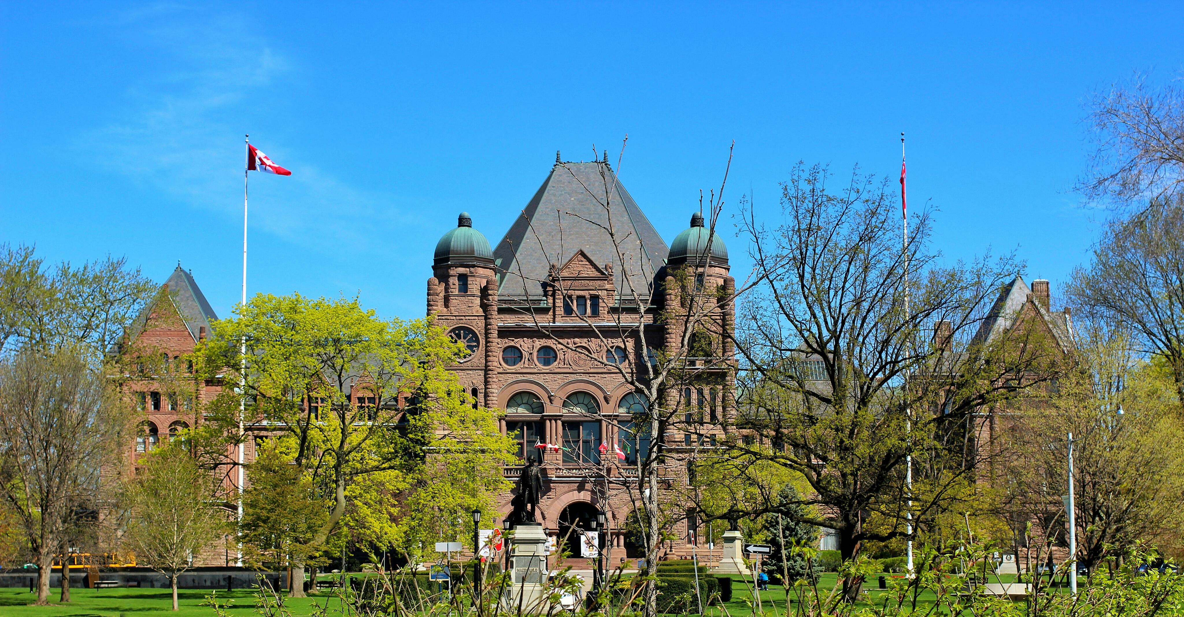 Historic building with a clock tower and Canadian flags, surrounded by lush green trees under a bright blue sky.