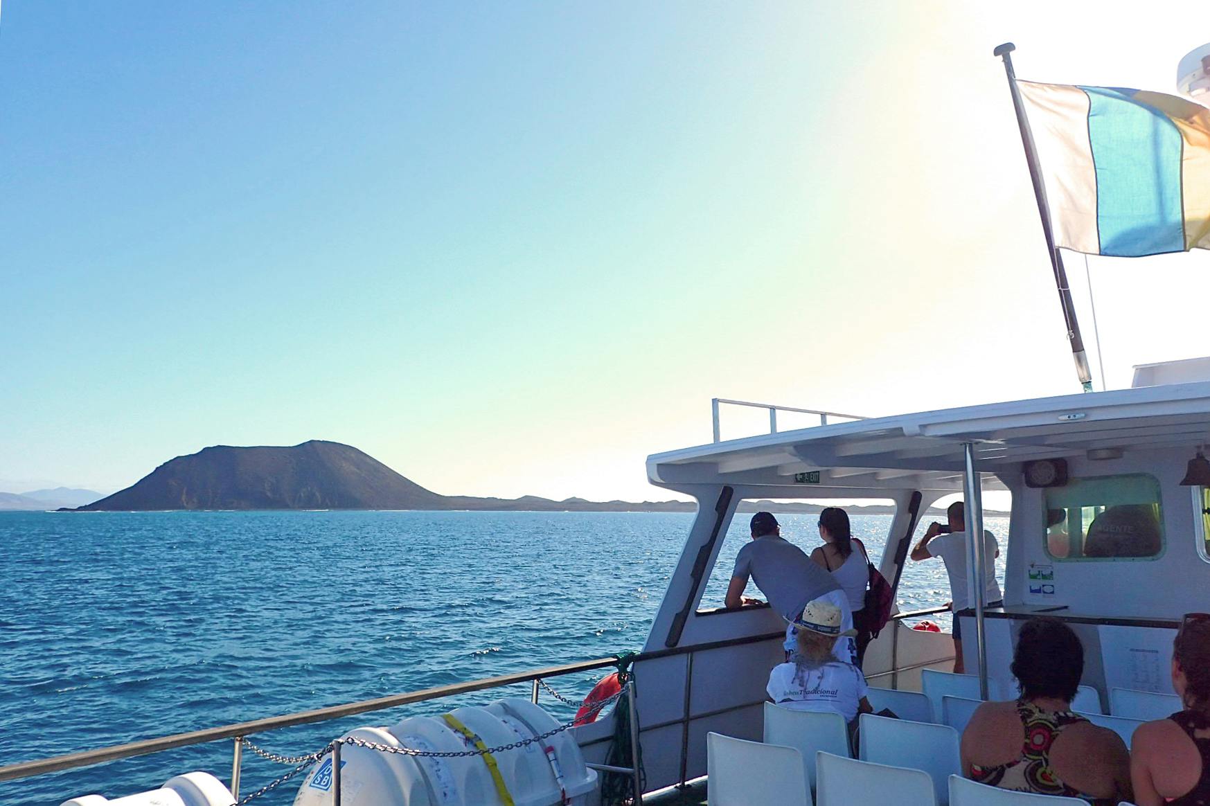 Several people on a boat looking at a distant island under a clear sky with a flag waving at the back.