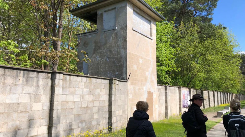 Two people in dark jackets walk near a tall, weathered guard tower beside a concrete wall, surrounded by trees and greenery.