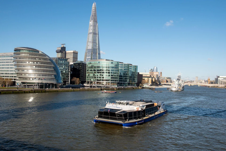 A boat on a river with modern buildings and the Shard skyscraper in the background, under a clear blue sky.
