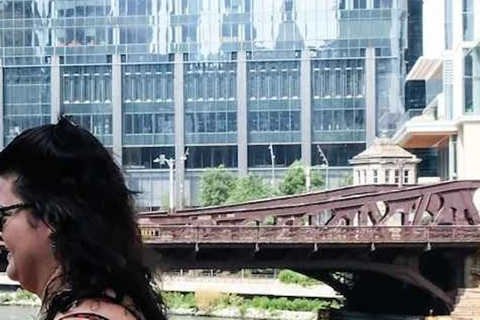 People walking on a bridge over a river with skyscrapers and a tour boat in the background on a sunny day.