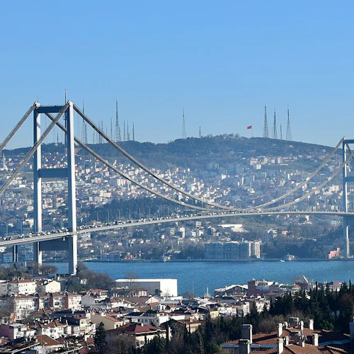 Hängbro som sträcker sig över en vattenkälla med en stadsbild och kullar i bakgrunden under en klarblå himmel.