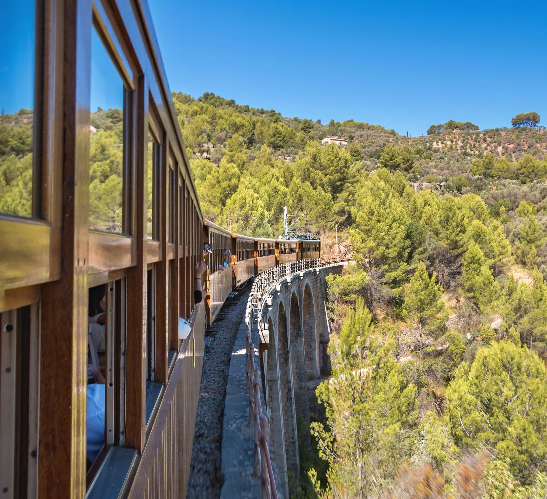 A train travels over a stone viaduct in a forested mountainous area under a clear blue sky.