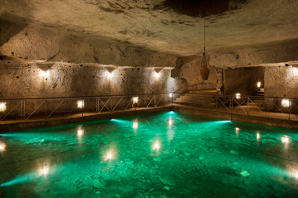 Underground room with illuminated green water pool, stone walls, steps, and hanging pottery. Fence with lights surrounds the pool.