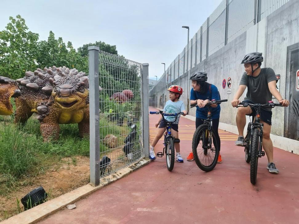 A child and two adults on bicycles pause near a fence with dinosaur models visible behind it on a paved pathway.