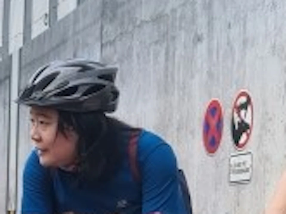 A child and two adults on bicycles pause near a fence with dinosaur models visible behind it on a paved pathway.