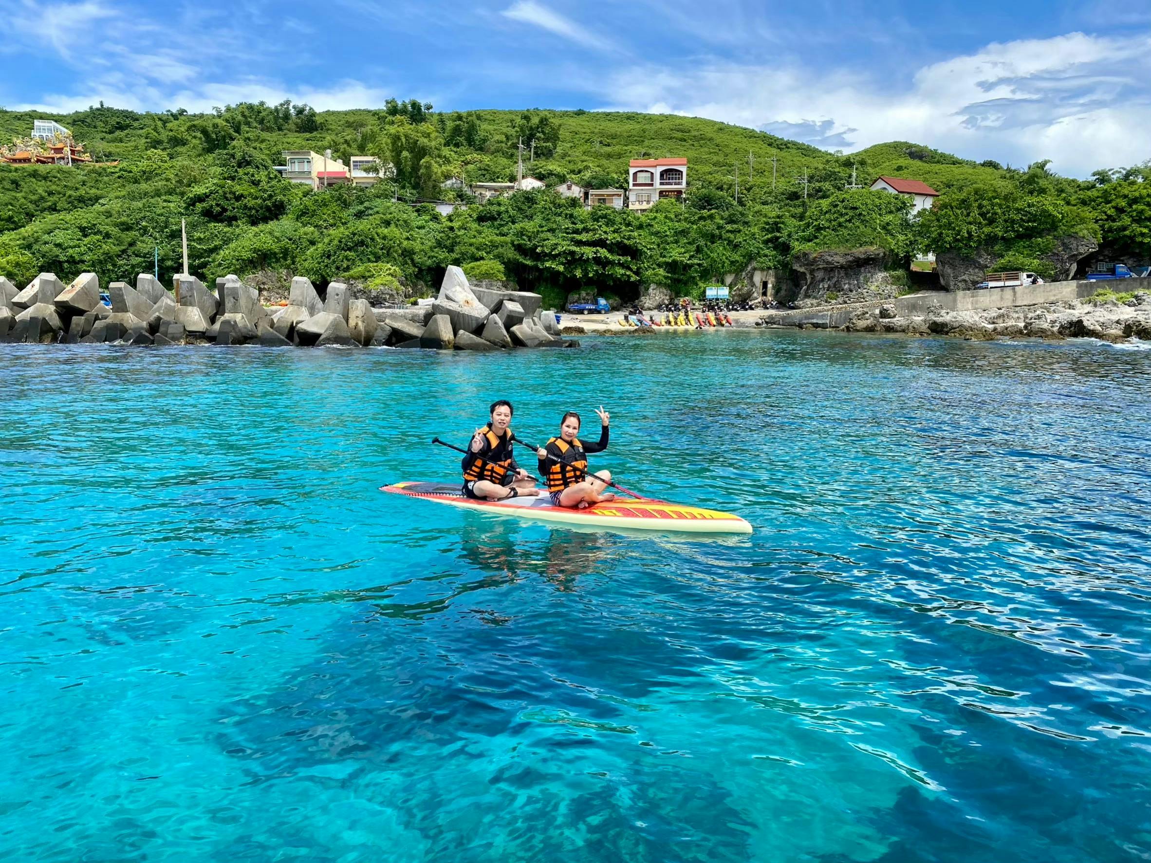 Two people wearing life vests sitting on a paddleboard in clear turquoise water near a rocky shoreline with greenery.