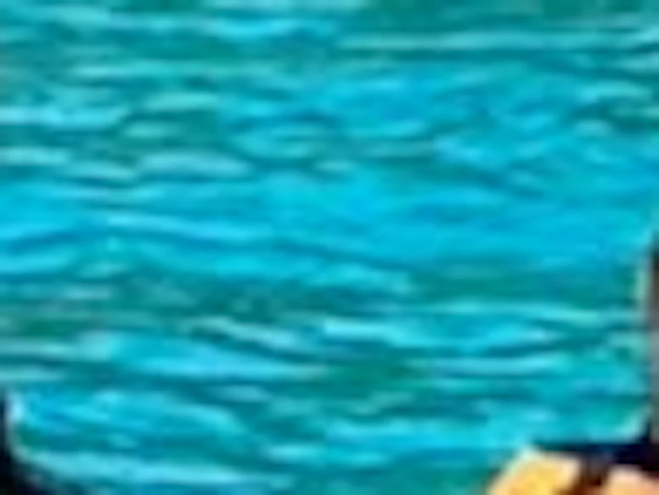 Two people wearing life vests sitting on a paddleboard in clear turquoise water near a rocky shoreline with greenery.
