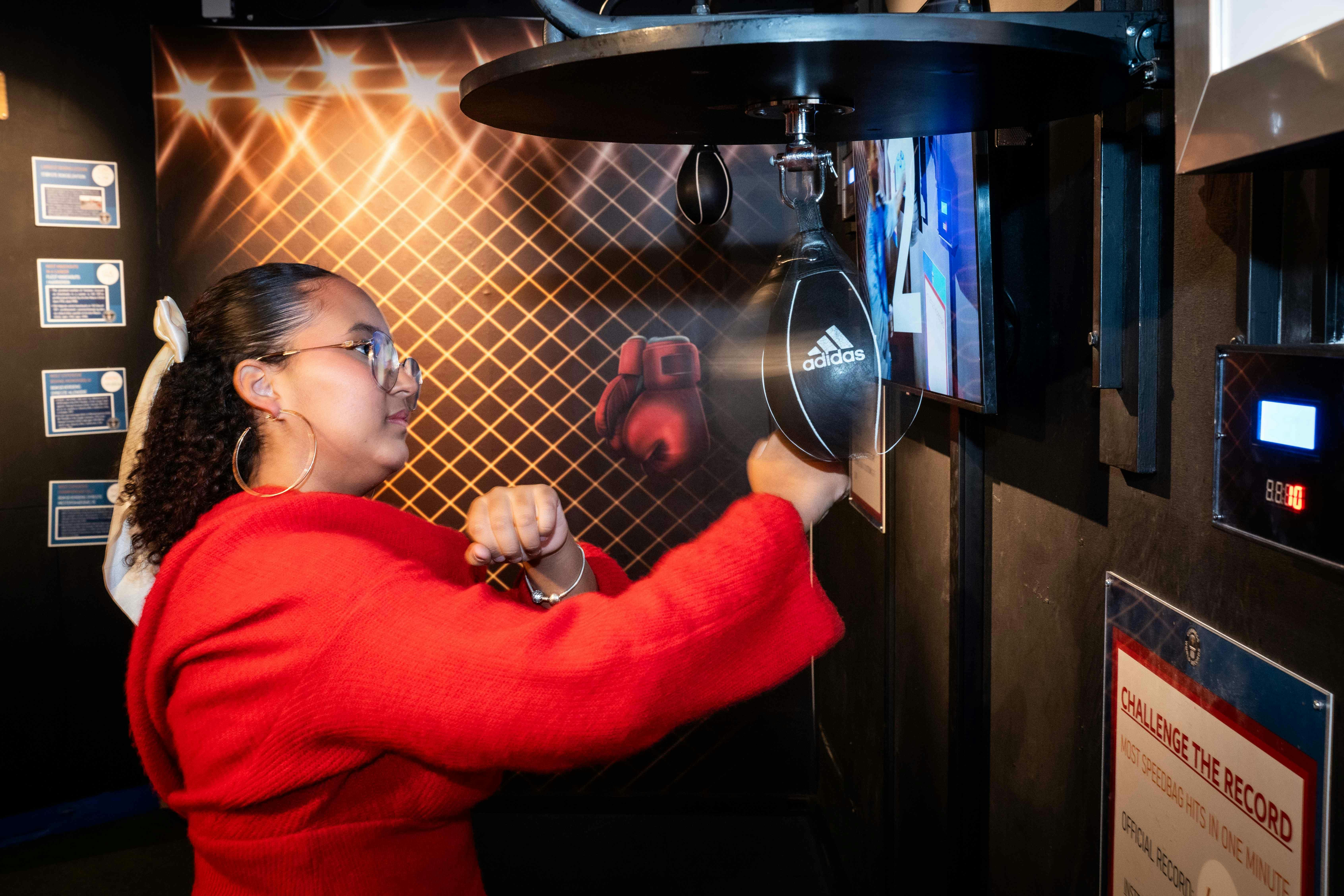 A person in a red sweater punches a speed bag in a sports training area with boxing gloves hanging in the background.