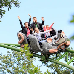 Fyra personer med upphöjda armar som rider på en berg- och dalbana formad som en mus, omgiven av grönska, på en klar dag.