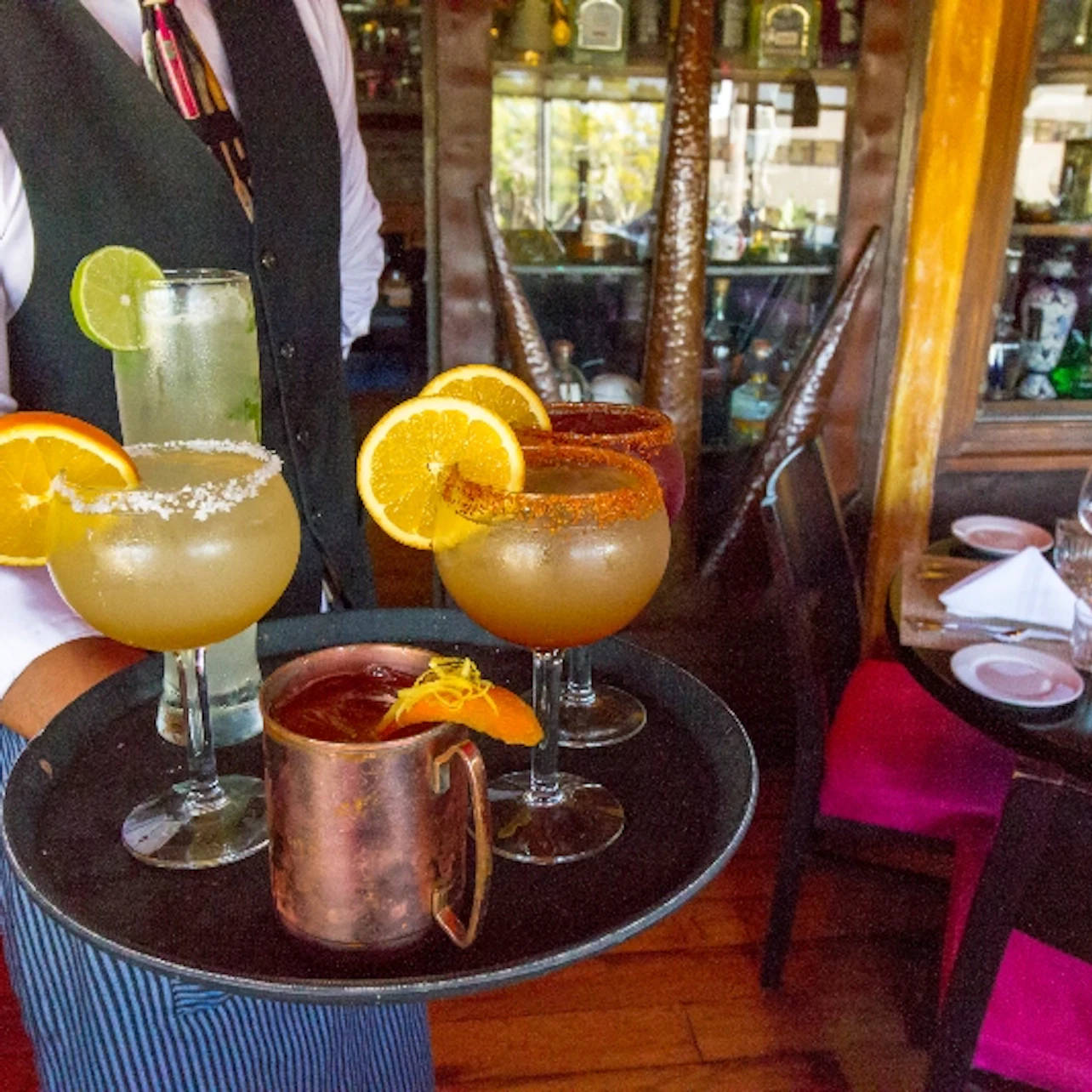 A waiter in a vest and tie holds a tray with several cocktails garnished with citrus slices in a restaurant setting.