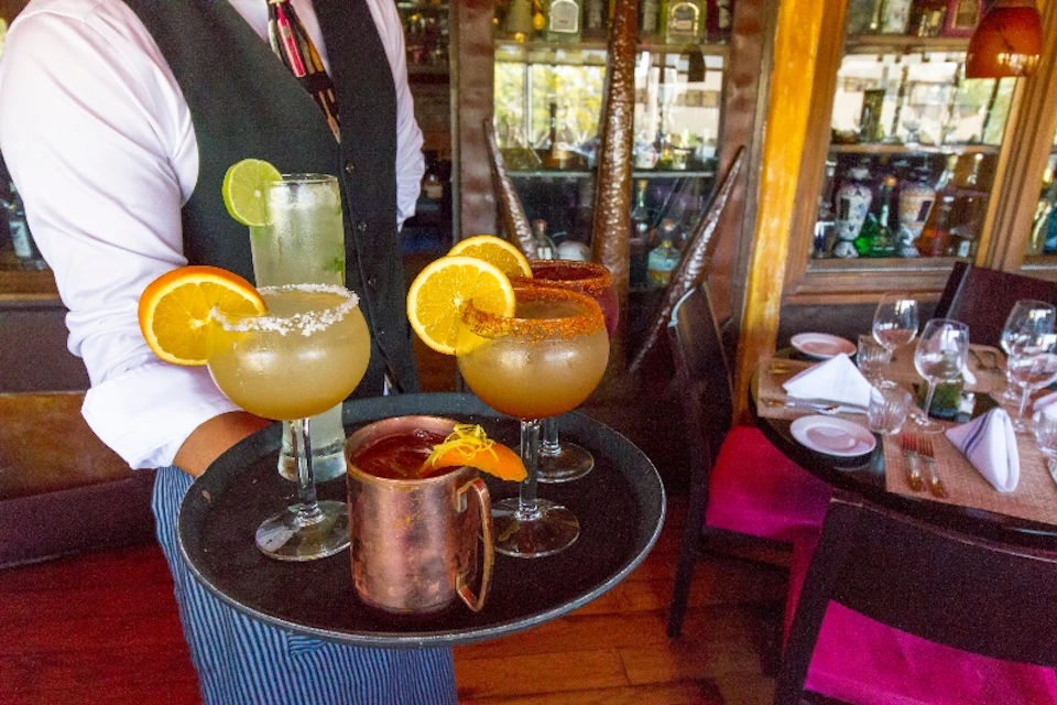 A waiter in a vest and tie holds a tray with several cocktails garnished with citrus slices in a restaurant setting.