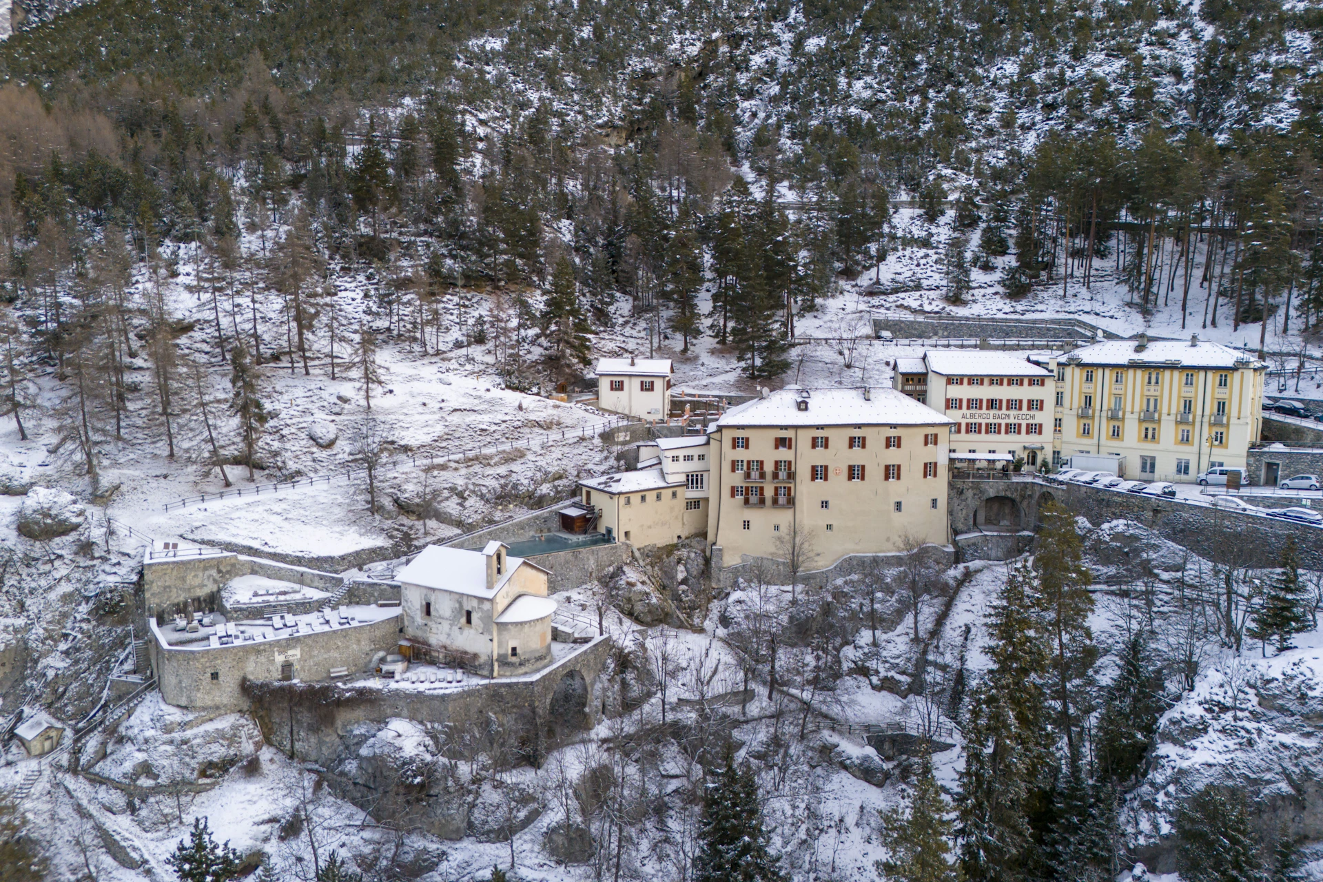 A snowy mountain village with several buildings, surrounded by trees and rocky terrain.