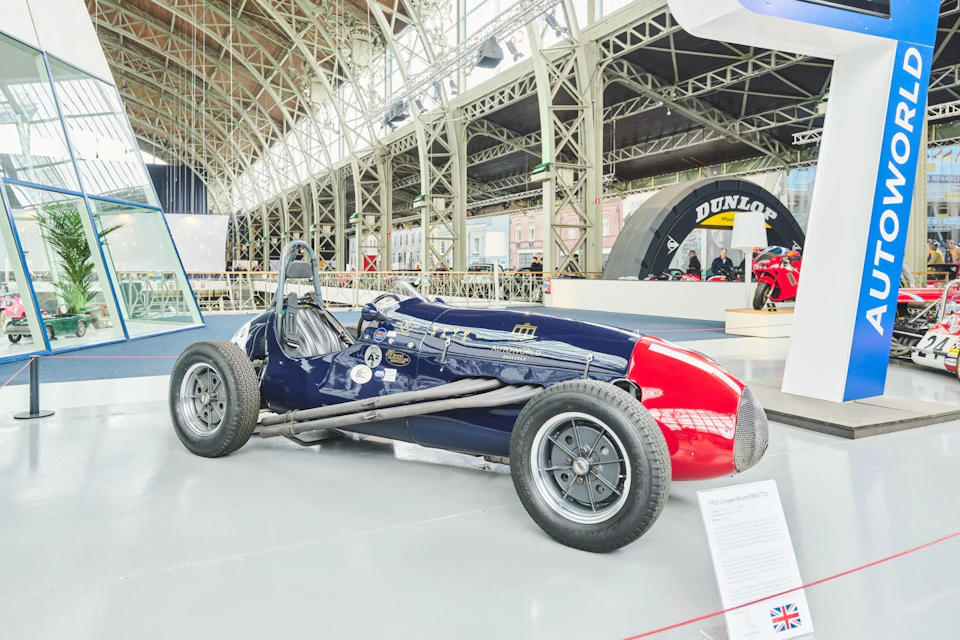 A vintage race car with a red front and blue body is displayed indoors on a smooth floor. Exhibition plaques and metal truss structure are visible.